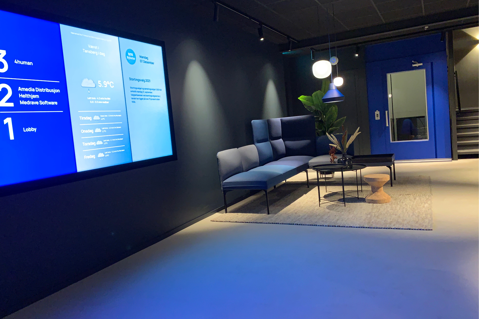 Lobby i kontorbygg med store skjermer, sofagruppe og knallblå heis